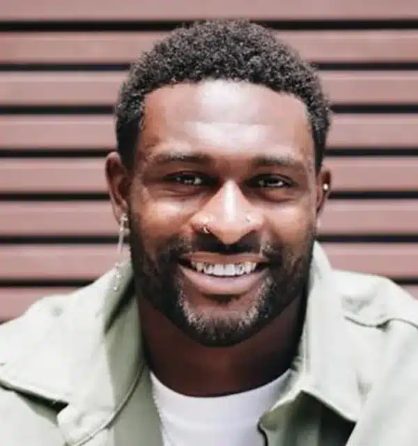 smiling man with short curly hair and earrings poses in front of a wooden backdrop.