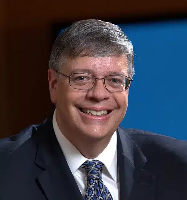 smiling man in glasses and a suit poses for a close-up portrait against a blurred background.