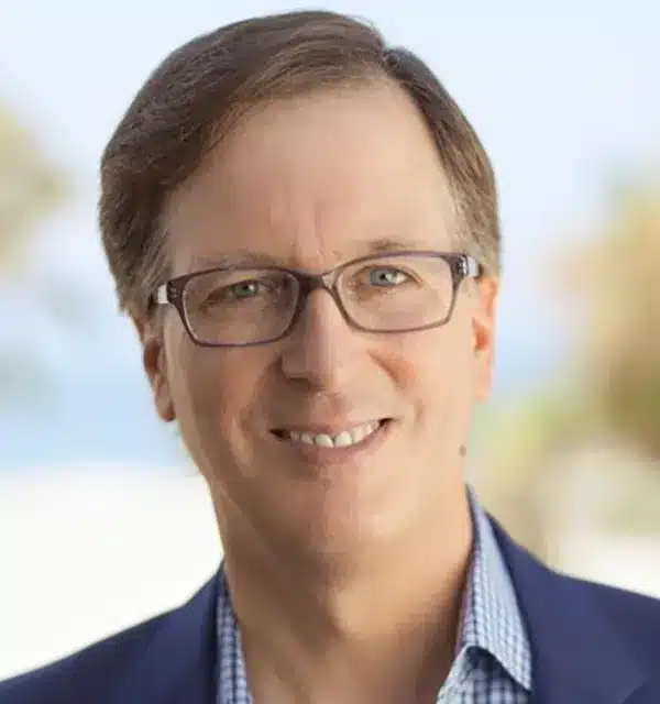smiling man with glasses and a blue blazer poses outdoors with a blurred background.
