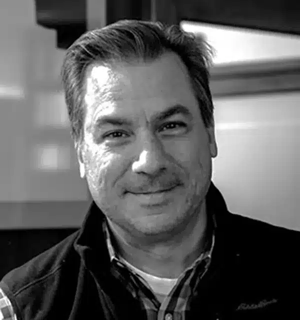 smiling man with short hair and a dark vest poses for a close-up photo against a blurred background.