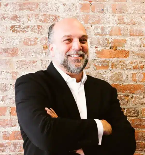 smiling man with a beard stands with arms crossed against a textured brick wall.