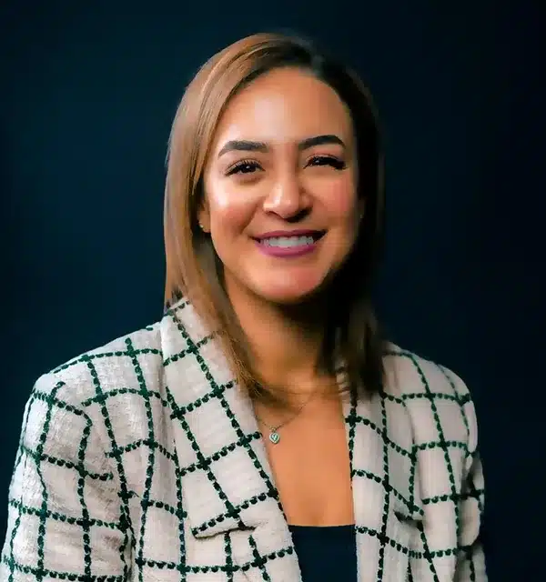 woman with shoulder-length hair smiles, wearing a patterned blazer against a dark background.