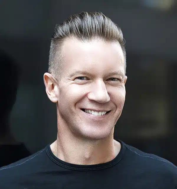 smiling man with short hair and a black shirt is photographed against a blurred background.