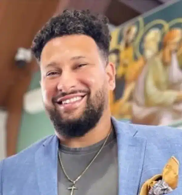 Smiling man in a blue blazer with a cross necklace, set against a colorful background.