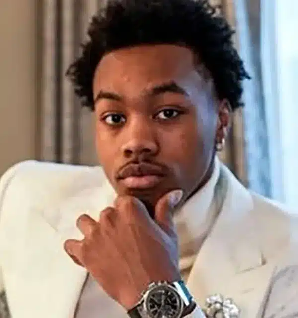 young man in a white suit poses thoughtfully, resting his chin on his hand, with a watch on his wrist.