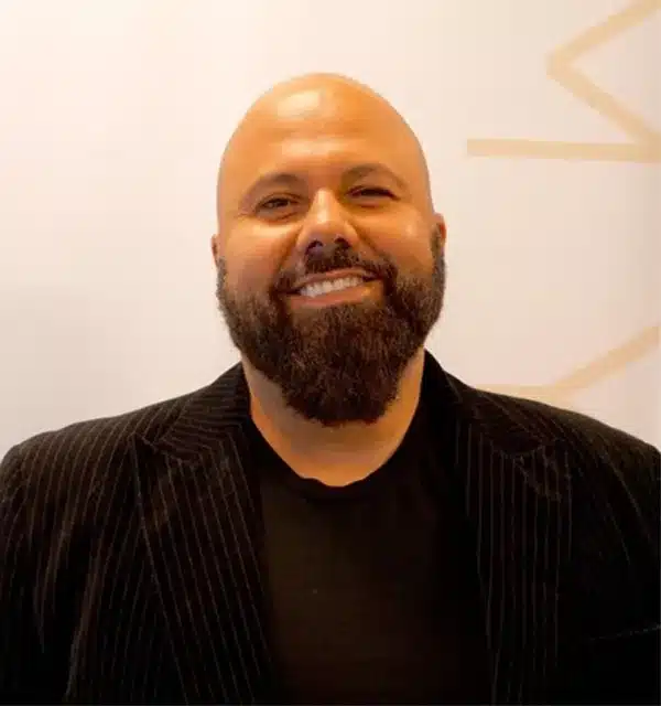 smiling man with a bald head and a beard wears a black jacket over a black shirt against a light background.